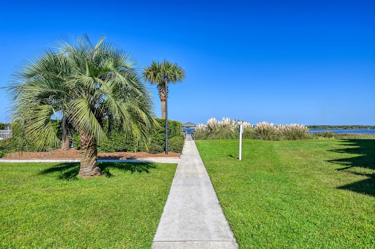 A scenic exterior shot featuring palm trees, a lush pathway, and tranquil water views. The pathway leads towards the water, framed by greenery and open sky, evoking an inviting, serene mood. The image suggests easy access to the water for walks or launching water activities and hints at the natural beauty of the area.