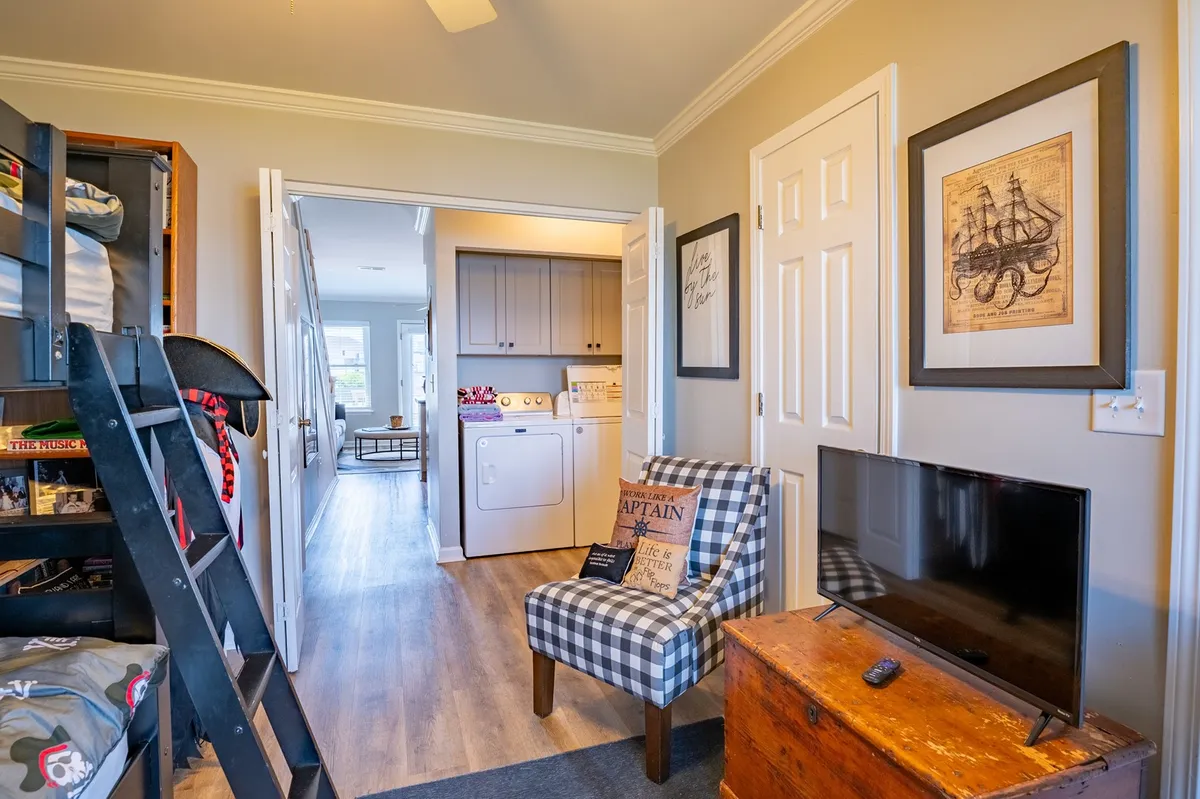 A functional and inviting laundry area featuring a plaid chair, washer/dryer unit, checkered throw pillow, television, and a wooden chest. The space doubles as a spot for relaxing or multitasking while handling chores.