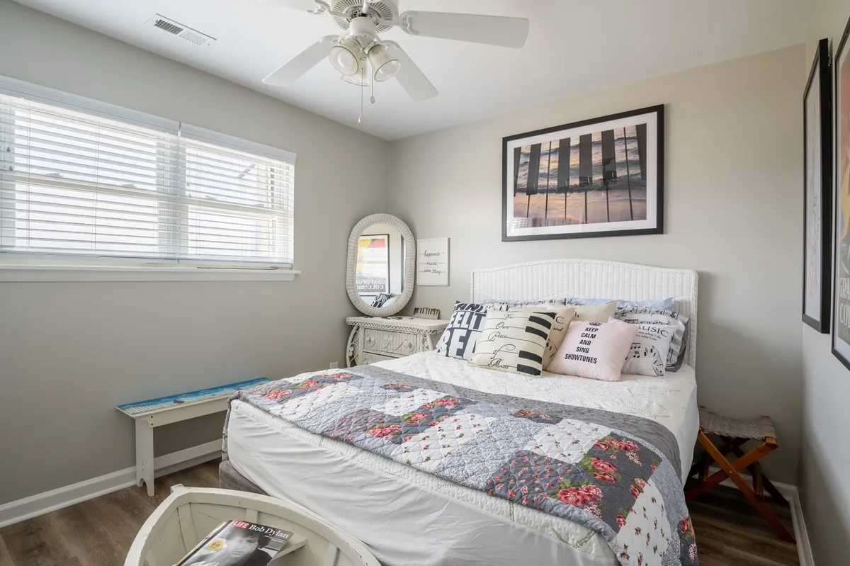A well-appointed bedroom with a bed adorned with decorative pillows, framed artwork, a round mirror, and a wooden bench. The space feels relaxed and comfortable, with soft tones and thoughtful decor.