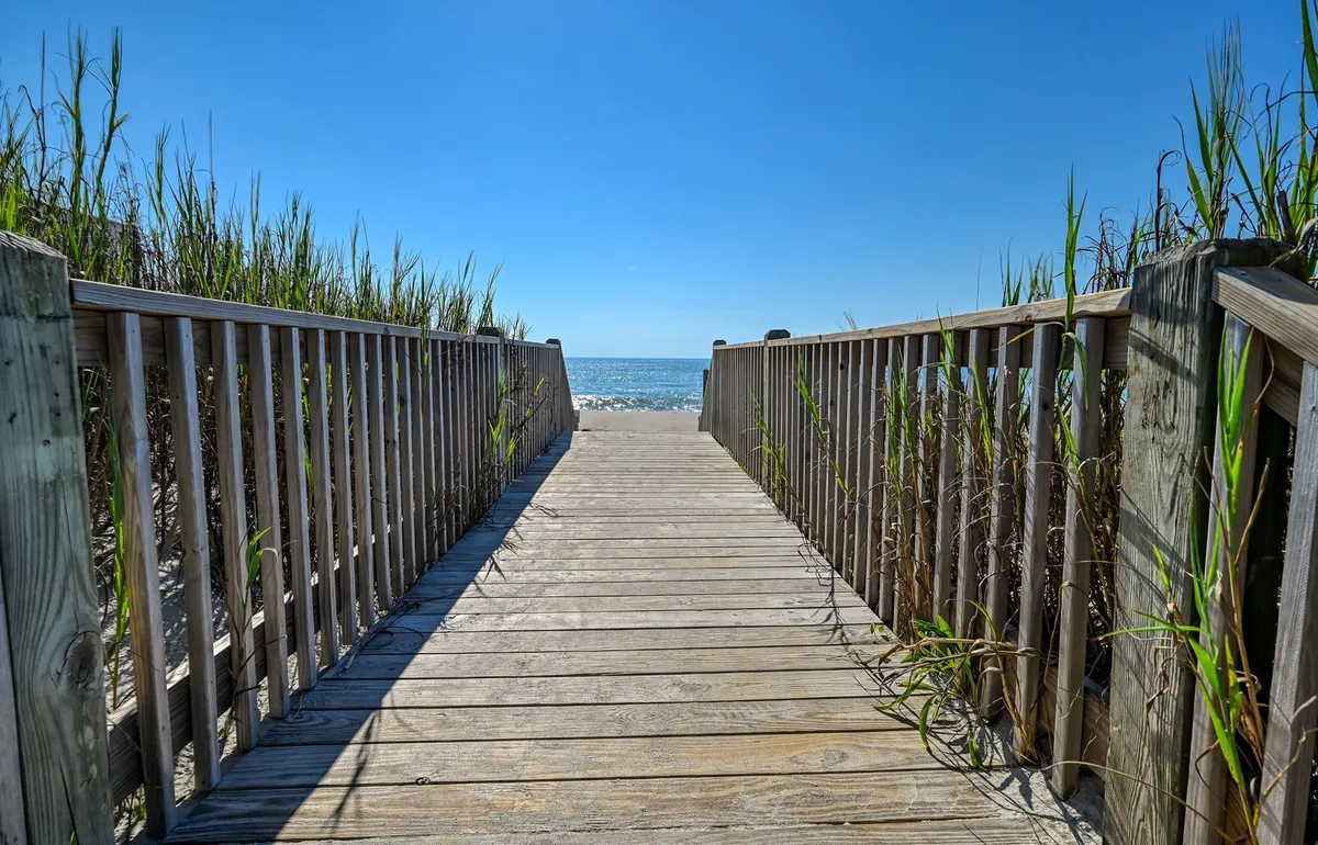 A wooden walkway bordered by dune grass leads out towards a panoramic view of the ocean. The scene captures the open sky, the gentle curve of the dunes, and the beach stretching into the distance, suggesting easy access for sunset strolls or peaceful mornings by the water.