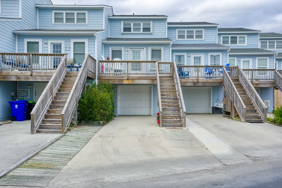 This image showcases the exterior of the Jolly Roger property, with sturdy wooden stairs leading up to townhome-style balconies furnished with outdoor chairs. Parked vehicles and a blue garbage can suggest a practical, lived-in community, while groups can be imagined enjoying the communal spaces. The scene captures the sense of a relaxed, social, and approachable environment.