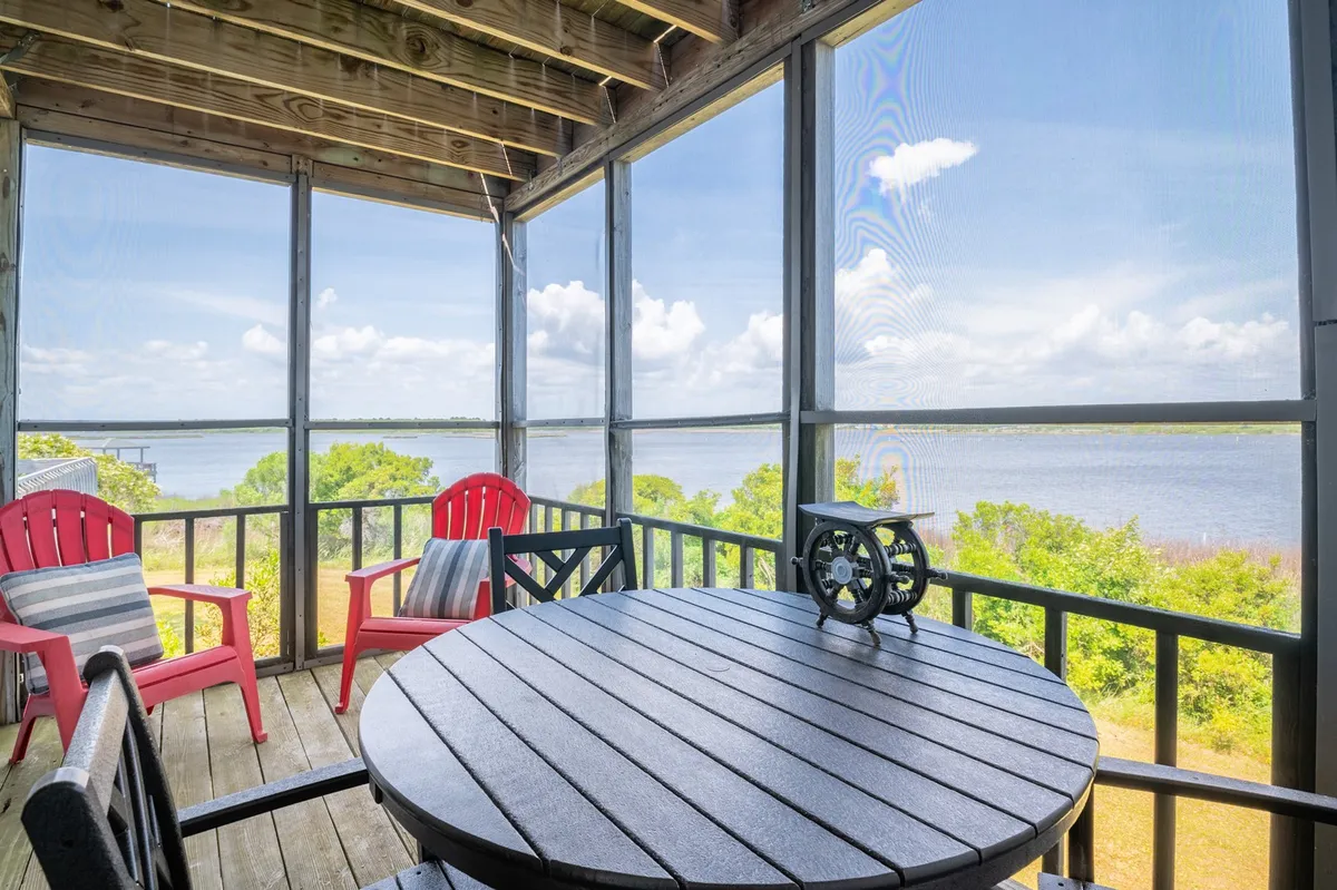 An exterior scene showing a wooden table accompanied by vibrant red chairs on a porch or deck, with a beautiful water view in the background. The setup suggests a welcoming place for dining, gathering, and overlooking the marsh or sound, under open skies.