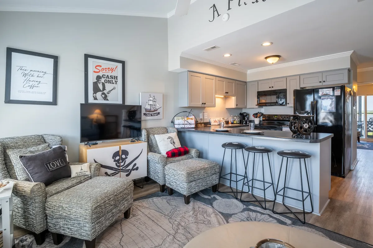 A welcoming open-concept living space featuring comfortable armchairs, a kitchen island with bar stools, decorative wall art, and a flat-screen television. The area is well-lit and designed for both relaxation and social gatherings, blending the living room and kitchen seamlessly.