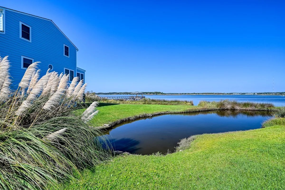 The image features the blue Jolly Roger condo nestled beside a serene pond edged with tall grasses, with the calm water reflecting the soft glow of sunrise. The peaceful setting showcases the natural beauty of North Topsail Beach, inviting relaxation and outdoor enjoyment such as fishing, bird watching, and evening gatherings by the water.