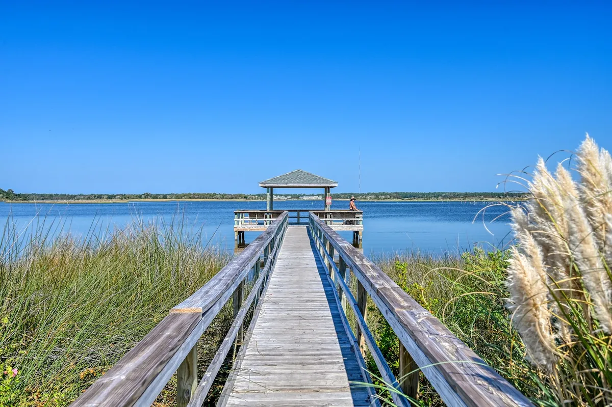 A long wooden pier stretches over calm water, leading to a charming gazebo at the end. The pier is framed by tall grasses, and the gazebo provides a shaded spot to enjoy the tranquil water views or try your hand at fishing. The overall mood is peaceful and inviting.