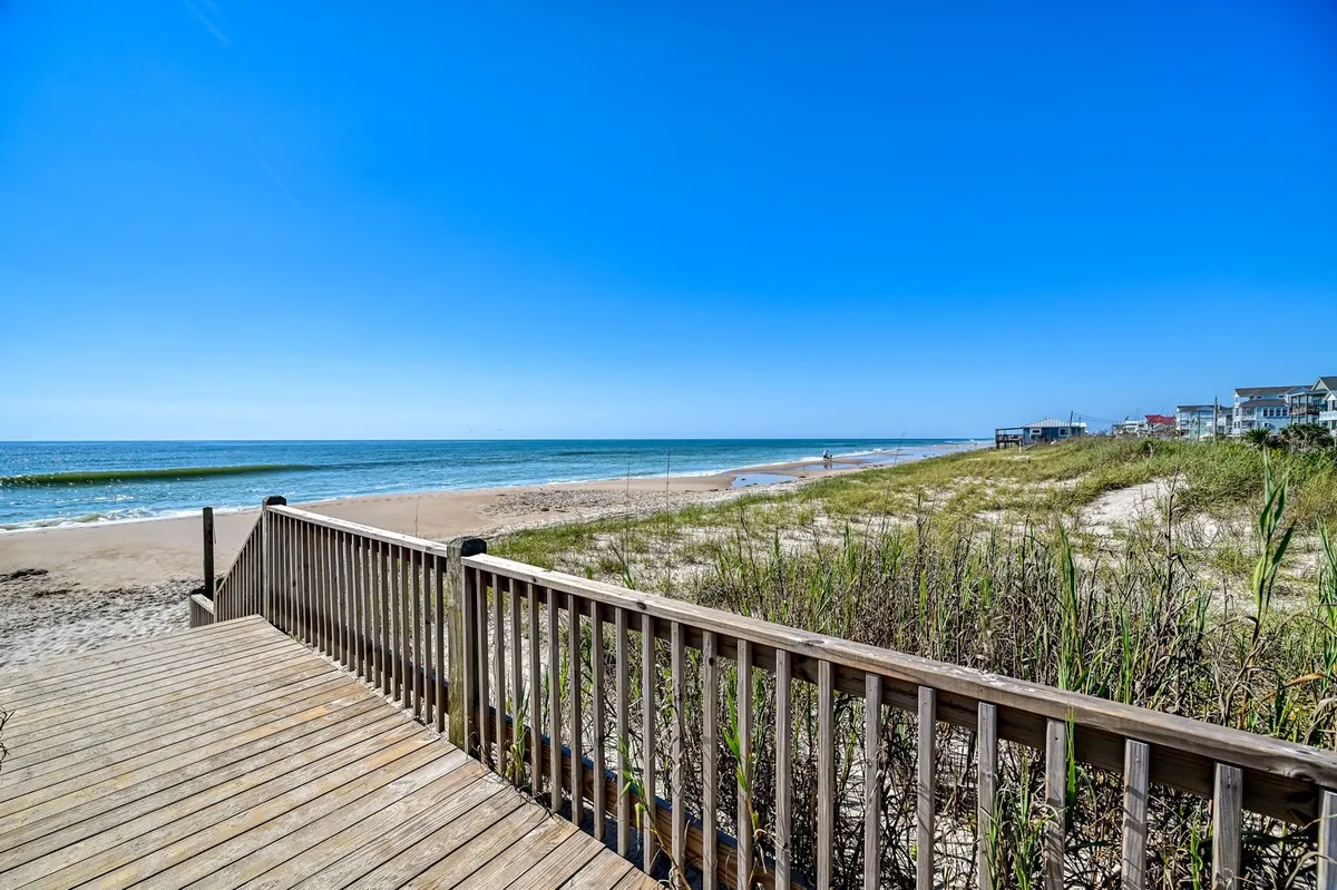 A scenic view from a wooden deck, overlooking a sandy beach with dune grass and the expansive ocean beyond. The relaxed mood is underscored by activities like sunbathing, fishing, and beachcombing. The image also hints at amenities like the fishing dock and soundfront pier mentioned in the section.