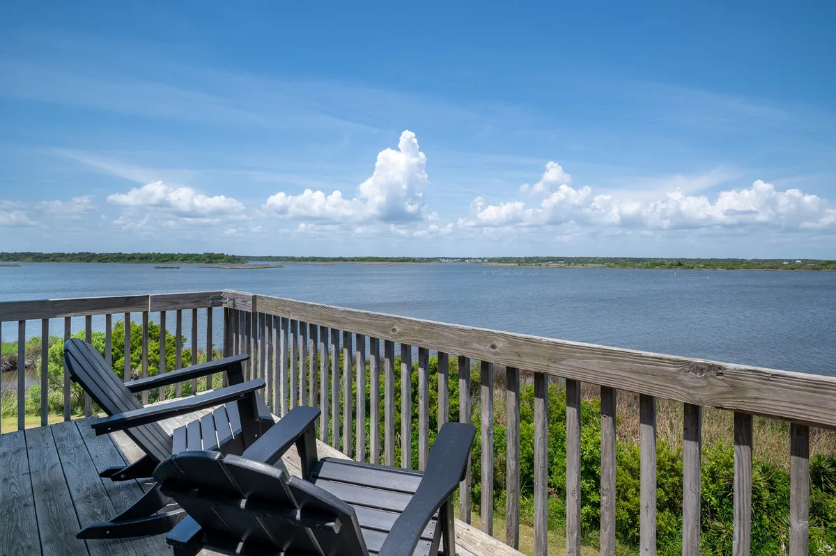 This image features a peaceful exterior scene with a wooden deck overlooking the water. Adirondack chairs are set up for relaxing and enjoying the view, with the sun low on the horizon, hinting at a beautiful sunset reflected in the water. The nearby soundfront access is clearly visible, inviting thoughts of launching a kayak or simply taking in the scenery.