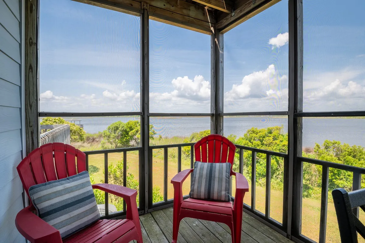 A screened-in porch featuring red Adirondack chairs positioned to overlook a scenic, tranquil view through the enclosing mesh. The porch is surrounded by natural greenery and hints at proximity to the water, evoking a sense of calm and relaxation. Soft lighting and the view outside make this space feel like a seamless extension of the interior, perfect for enjoying sunsets and observing wildlife.